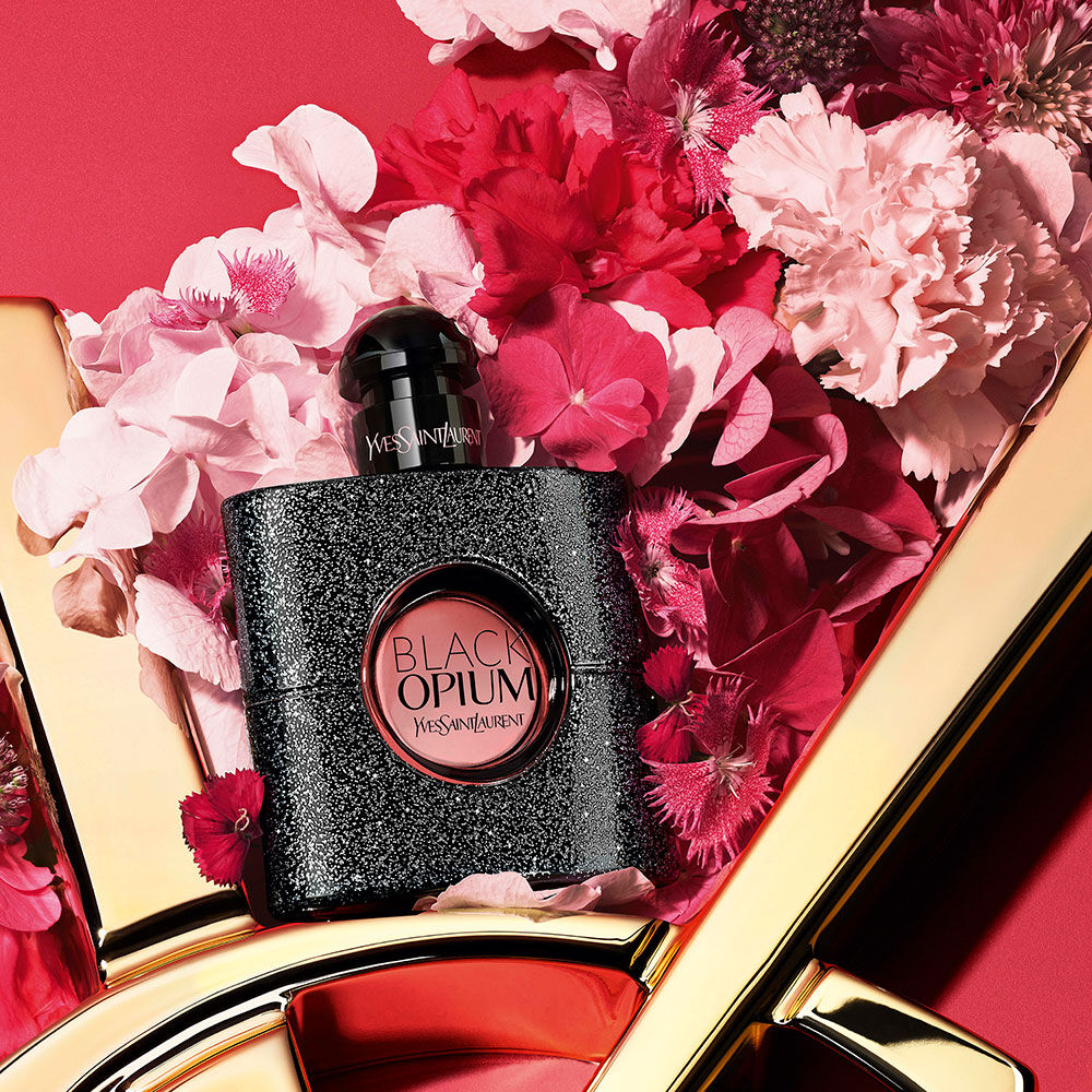 Black Opium Yves Saint Laurent glittered black perfume bottle surrounded by pink and red flowers, styled on a red background with a gold YSL logo detail.