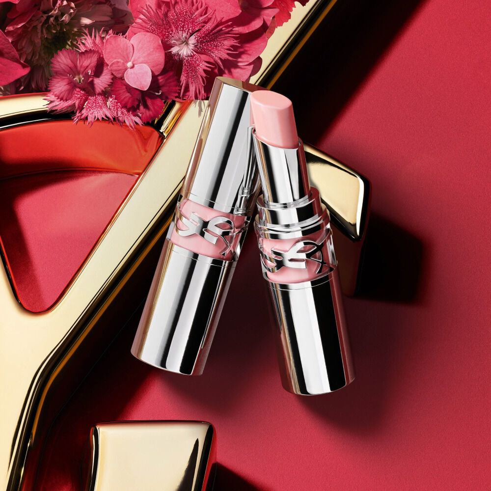 YSL Loveshine lipstick shade 44 in silver packaging, shown open with a soft pink bullet beside the closed tube, styled with pink flowers on a red background with gold YSL logo detail.