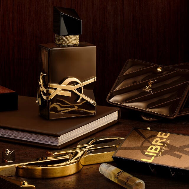 Libre Santal Couture bottle on a desk with a YSL leather pouch, a notebook, and a gold mirror.