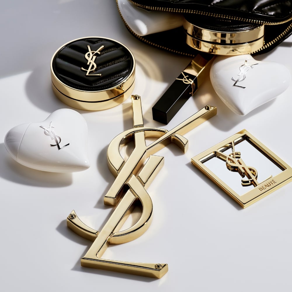 YSL makeup and Care Crush products spilling from a black bag around a gold Cassandre logo. Gold & black lifestyle.